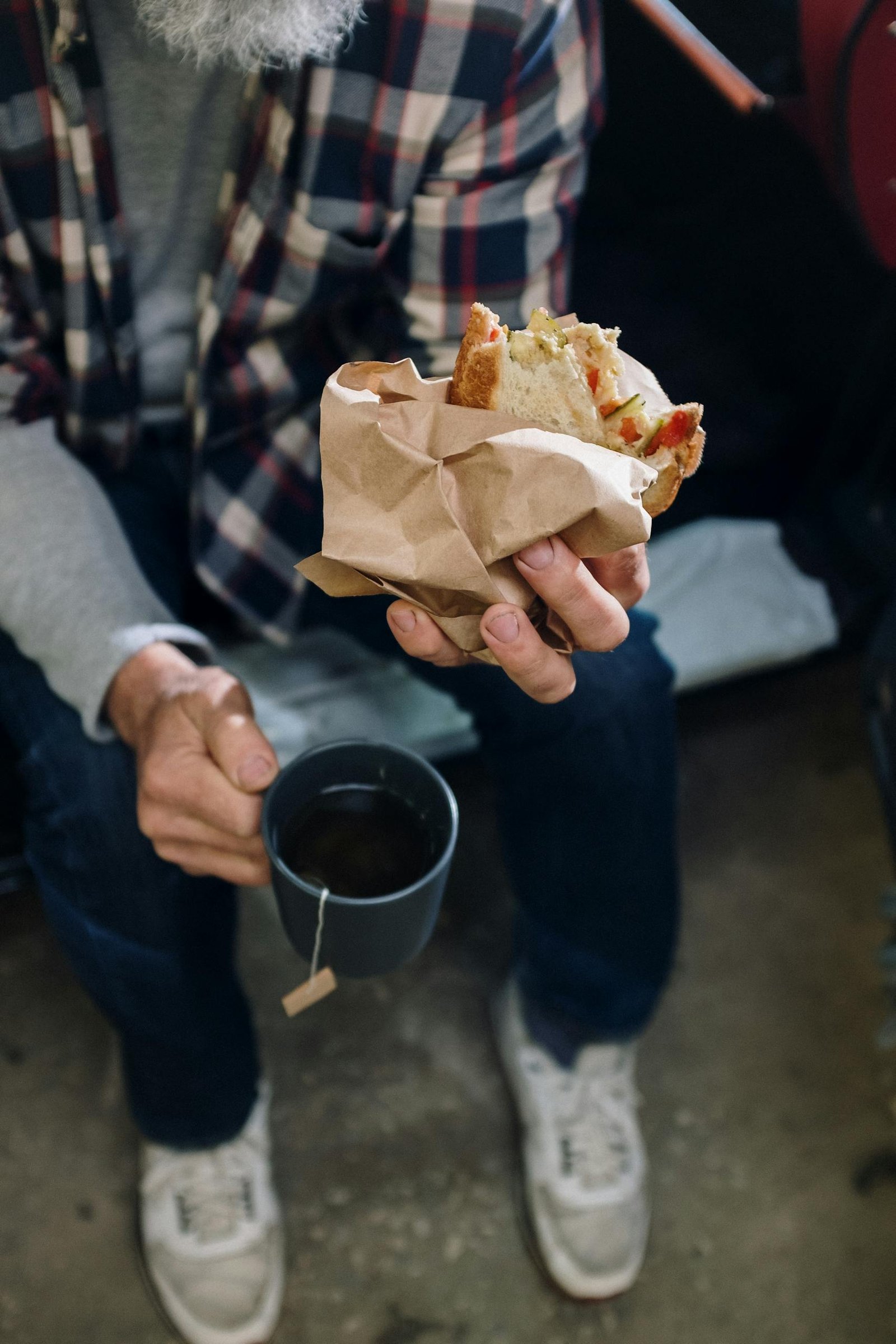 An adult man in casual attire holding a sandwich and a mug of tea, seated indoors.